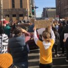 People at an intersection hold signs saying “Black Lives Matter” and “No Police State”.