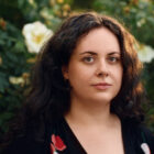 A white woman poses in front of some bushes with white flowers.
