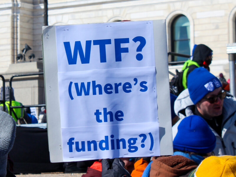 A protester holds a sign saying “WTF? (Where’s the funding?)”.