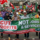 A crowd of marchers holds a banner reading "Cut profits, not jobs & services, support our transport workers".