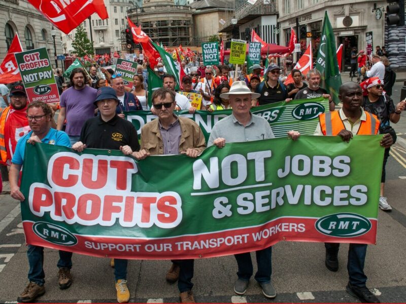 A crowd of marchers holds a banner reading "Cut profits, not jobs & services, support our transport workers".