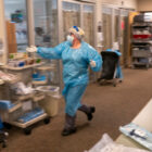 A person in blue PPE suit and white mask, gloves, and face shield rushes down a hallway reaching out a hand to a hand sanitizer.