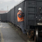A man in a white hard hat and orange safety vest hangs on the side of a black train freight.