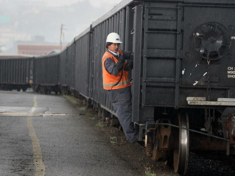 A man in a white hard hat and orange safety vest hangs on the side of a black train freight.
