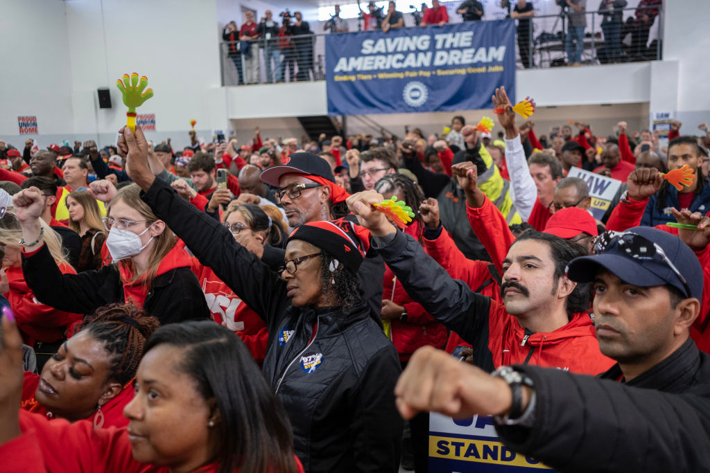 A crowd in red attire raises their fists, many with eyes closed, with a sign saying “Save the American Dream” behind them.