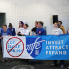 A dozen people hold a banner saying "MFE: Invest, Attract, Expand" and signs saying "MPS: PAY Us What You OWE Us!".