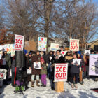 A woman speaks at an outdoor podium surrounded by signs reading "Neighbors Say ICE Out!" and "Stay Out of Our Schools.".