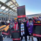 A crowd of people dressed in winter gear hold signs with names of people abducted by ICE walk outside near a airport terminal.