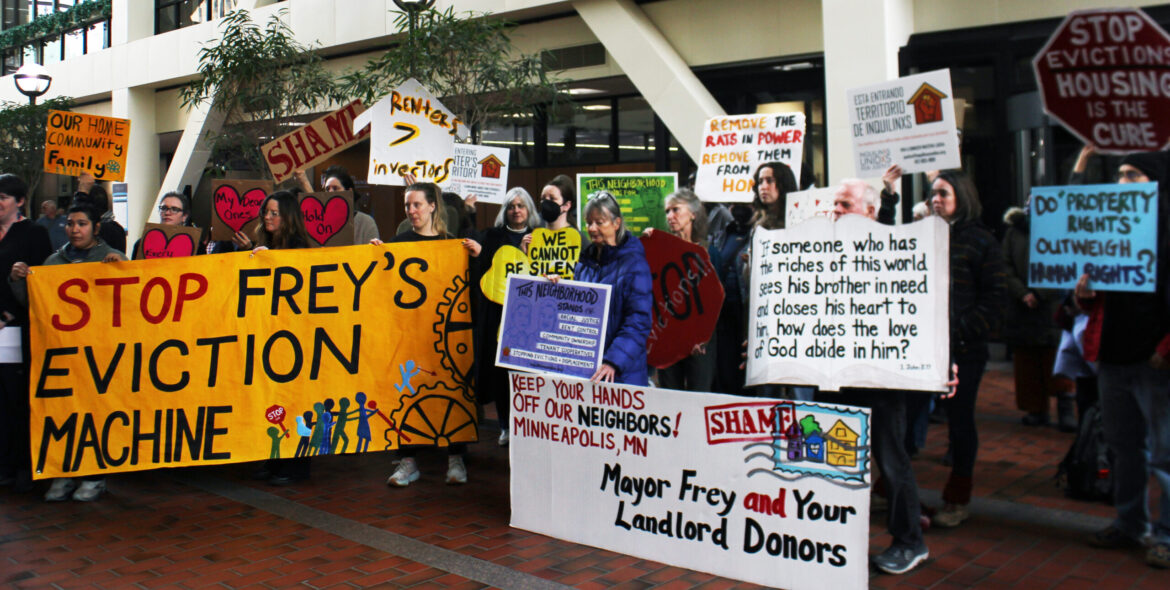 A crowd of people in a courtyard hold banners and signs saying “Stop Frey’s eviction machine” and “Our home, community, family”.