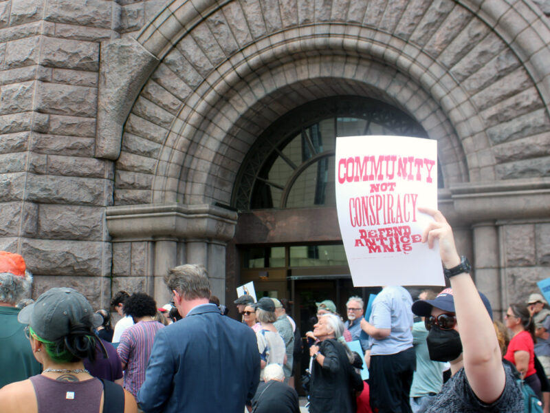A crowd of people facing away from the camera include a person wearing a black face mask and lifting up a sign that reads “community not conspiracy defend the anti ice MN 15” in red text